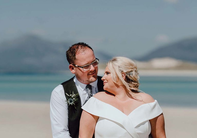 couple standing on beach with water behind them looking into each other's eyes in wedding dress and suit 